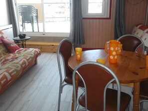 Ein gemütliches Zimmer mit Holzboden, einer Couch mit bunter Decke, einem Esstisch mit Stühlen und einem Fenster mit Vorhängen.