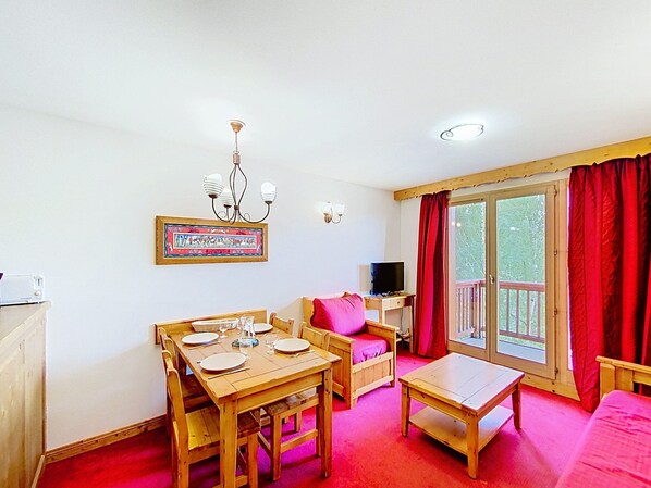 Une pièce chaleureuse avec une table à manger dressée pour quatre personnes, un canapé, une télévision et un balcon agrémenté de rideaux rouges.