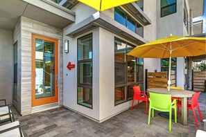 An outdoor seating area with a table and chairs, a yellow umbrella, and a building with large windows and an orange door. - Spectacular Living in Palm Springs' Coveted Vibe Community (Palm Springs)