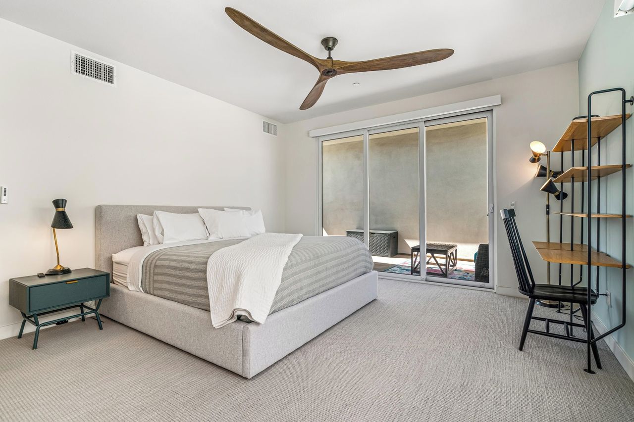 A bedroom with a bed, a nightstand, a chair, a ceiling fan, and a sliding glass door leading to a balcony.