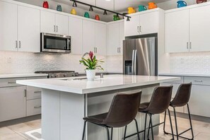 Una cocina moderna con armarios blancos, electrodomésticos de acero inoxidable, una isla central con taburetes de bar y detalles decorativos coloridos en los armarios superiores.