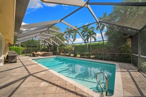 Piscina externa com área de estar coberta, cercada por palmeiras e céu claro.