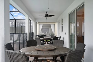 A covered patio with a round wooden table and wicker chairs, a ceiling fan, and a view of the backyard. - Stunning 9BR Villa Near Disney Storey Lake (Kissimmee)
