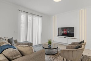 A modern living room with a flat-screen TV mounted on the wall, a round coffee table, and two armchairs. - Modern Home w/ Private Pool Near Disney (Kissimmee)