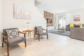 Een moderne woonkamer met twee grijze fauteuils, een klein houten bijzettafeltje en een grijze bank met gele kussens. Een trap leidt naar een hoger gelegen verdieping.