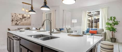 A modern kitchen with a central island, stainless steel appliances, pendant lighting, and a view into the living area.