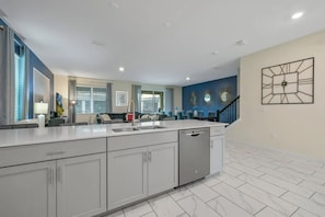 A modern kitchen with a central island, white cabinetry, and a dishwasher. There is a dining area with a table and chairs, and a staircase leading to another level. - Luxury 9BR Villa w/ Private Pool & Themes (Kissimmee)