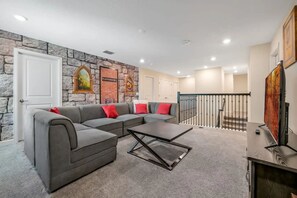 Un salon avec un canapé d’angle gris, une table basse et un téléviseur posé sur un meuble. On y trouve un mur en pierre orné de tableaux encadrés et un escalier avec une rampe.