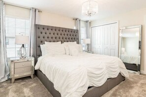 A bedroom with a tufted headboard, a large mirror, a bedside table with a lamp, and a carpeted floor. - Home w/ Private Pool 10 min to Disney (Kissimmee)