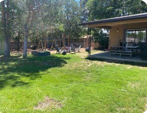 A backyard with a swing set, a covered patio area, and a lawn. - Modern Farmhouse in Uptown ABQ (Albuquerque)