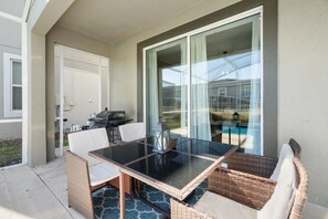 Eine überdachte Terrasse mit einem Glastisch, Korbstühlen, einem Grill und Blick auf einen Swimmingpool.