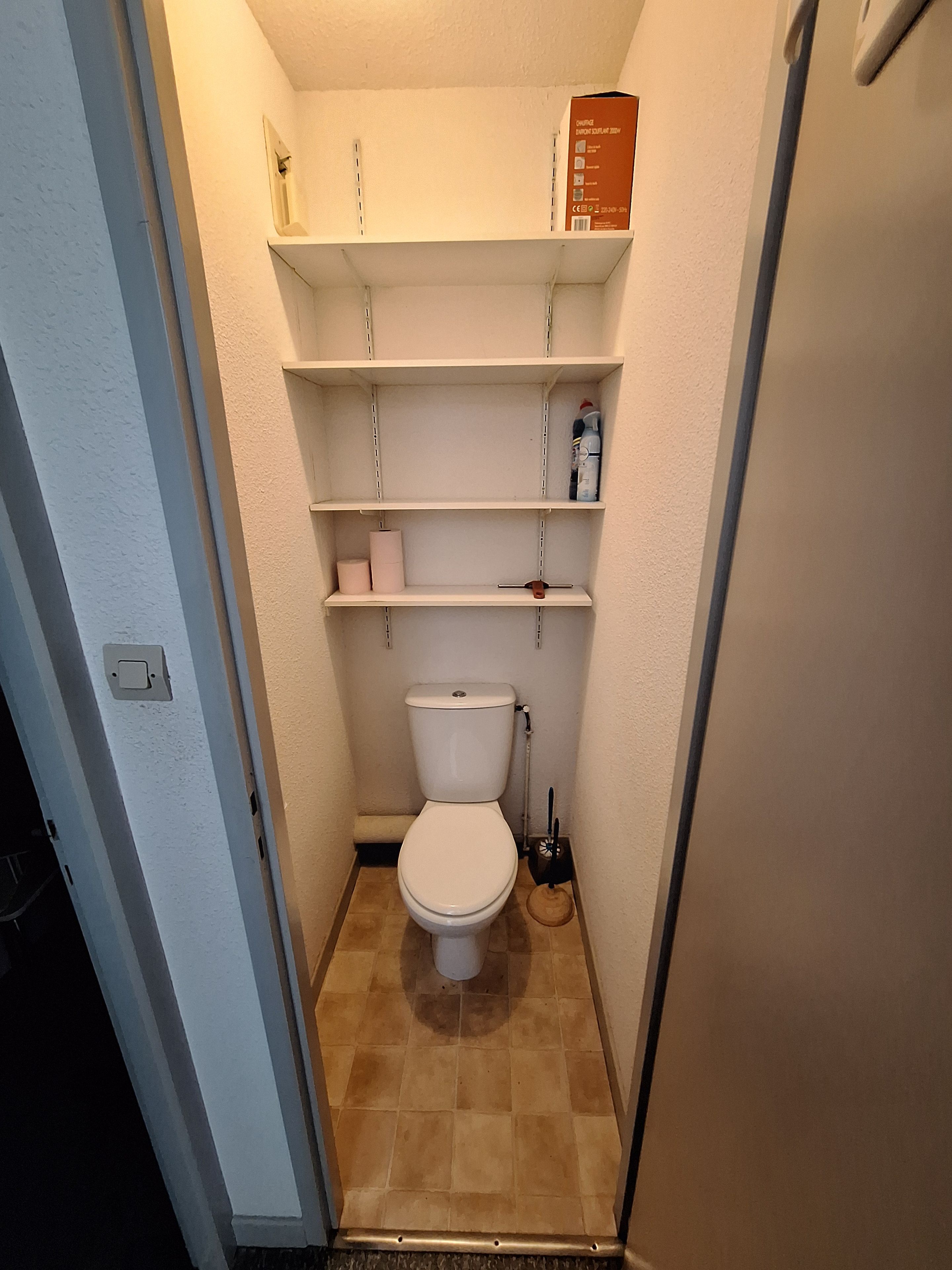 Une petite salle de bain étroite, avec des toilettes, des étagères et une boîte fixée au mur.