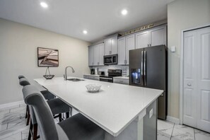 A modern kitchen with grey cabinets, a stainless steel refrigerator, a microwave, and a large island with bar stools. - Modern Home With Private Pool at Champions Gate (Davenport)