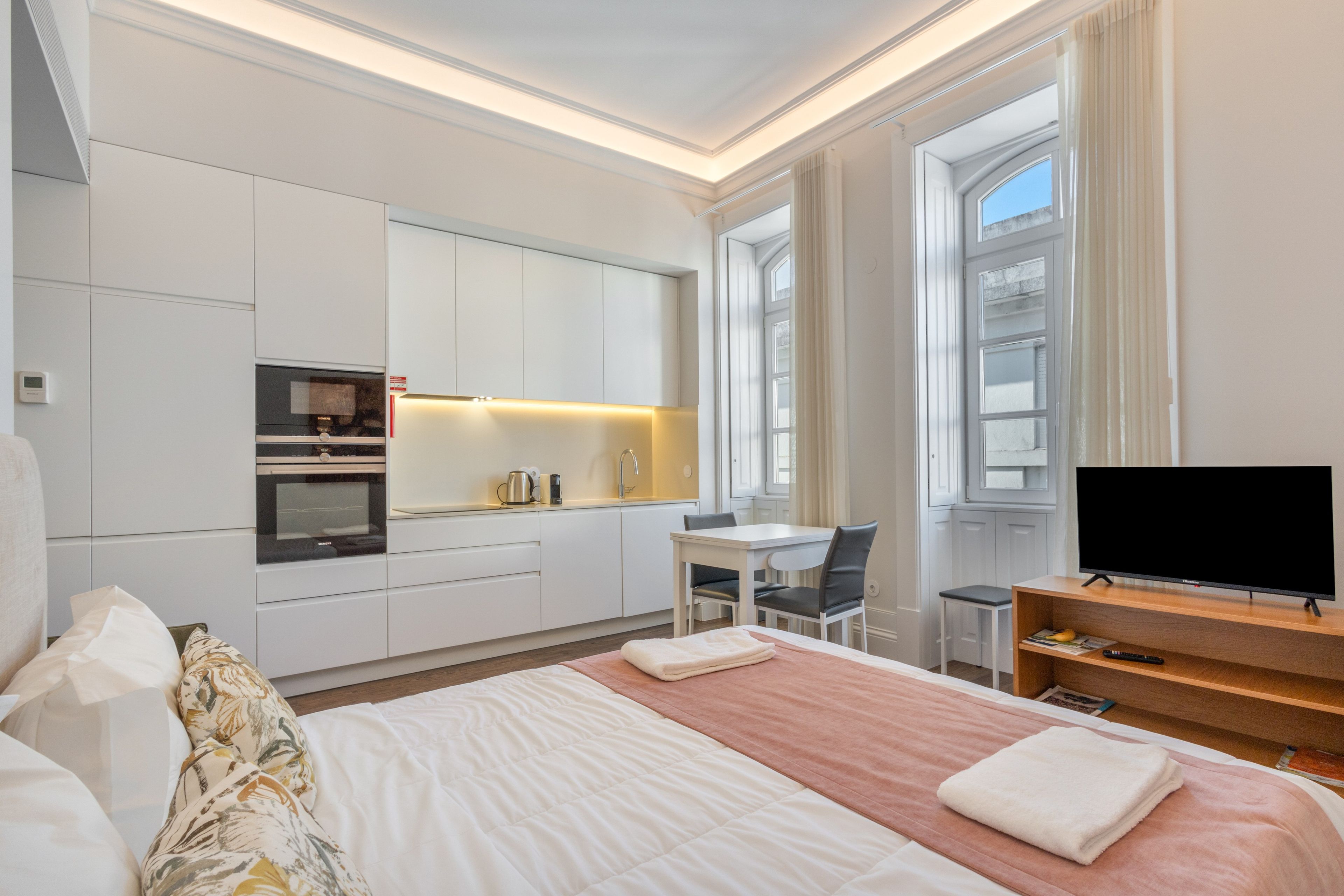 A modern hotel room with a bed, a desk, a television, and a kitchenette.
