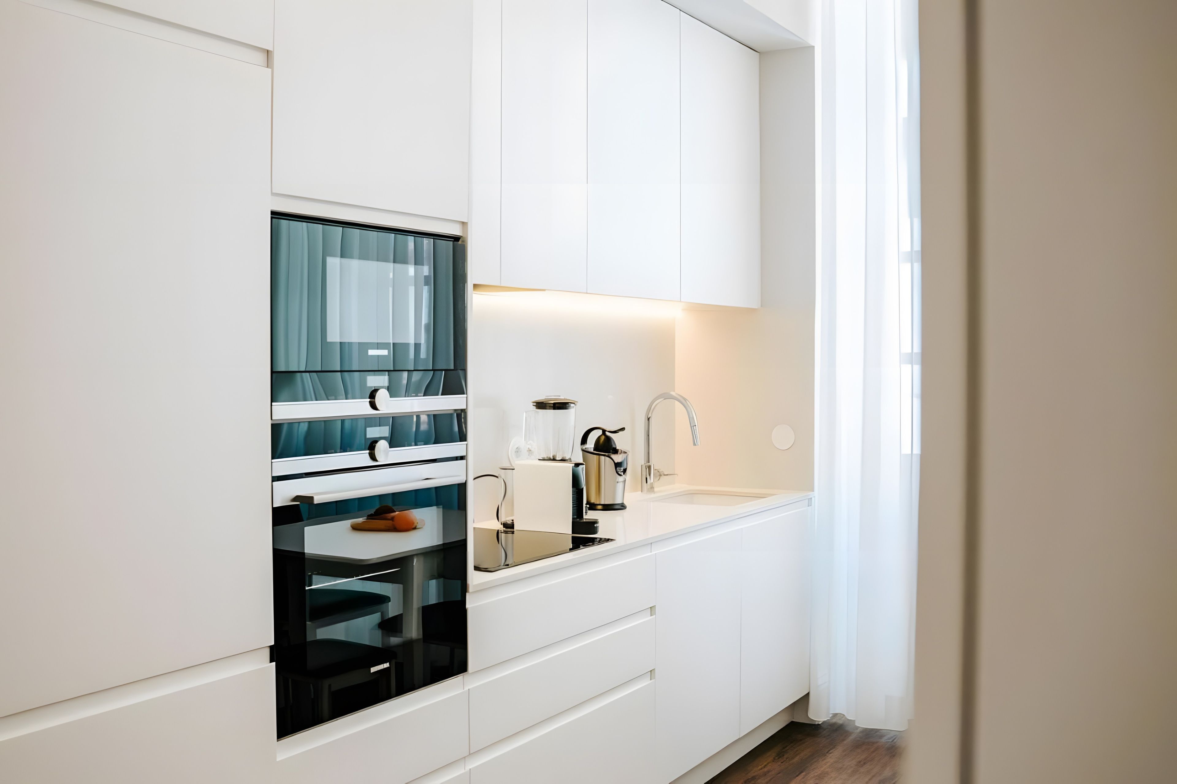 Una cocina moderna con armarios blancos, horno integrado, un lavamanos y una ventana con cortinas de gasa.