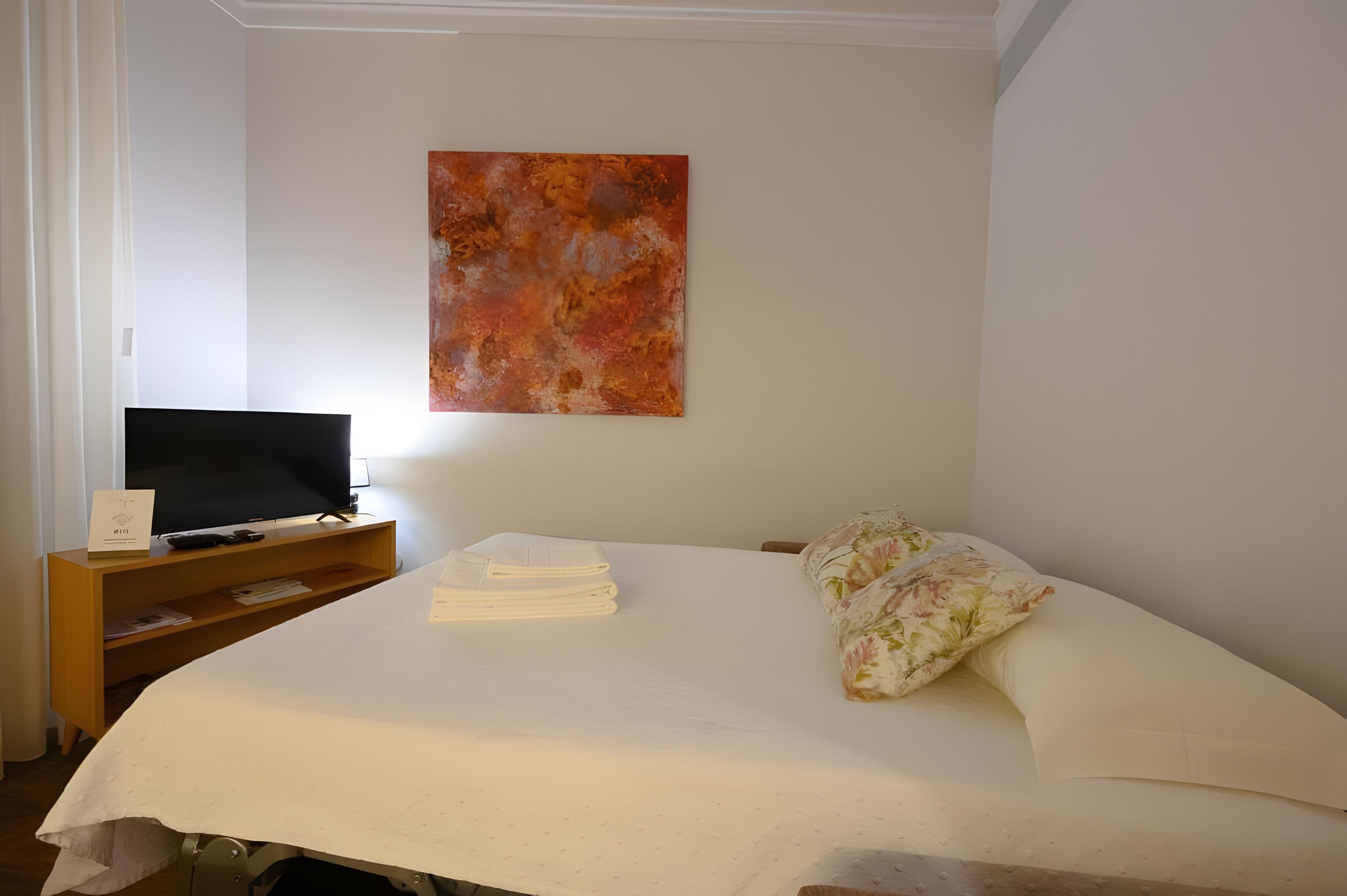 Een hotelkamer met een bed, een televisie op een houten standaard, een schilderij aan de muur en een bloemenkussen.