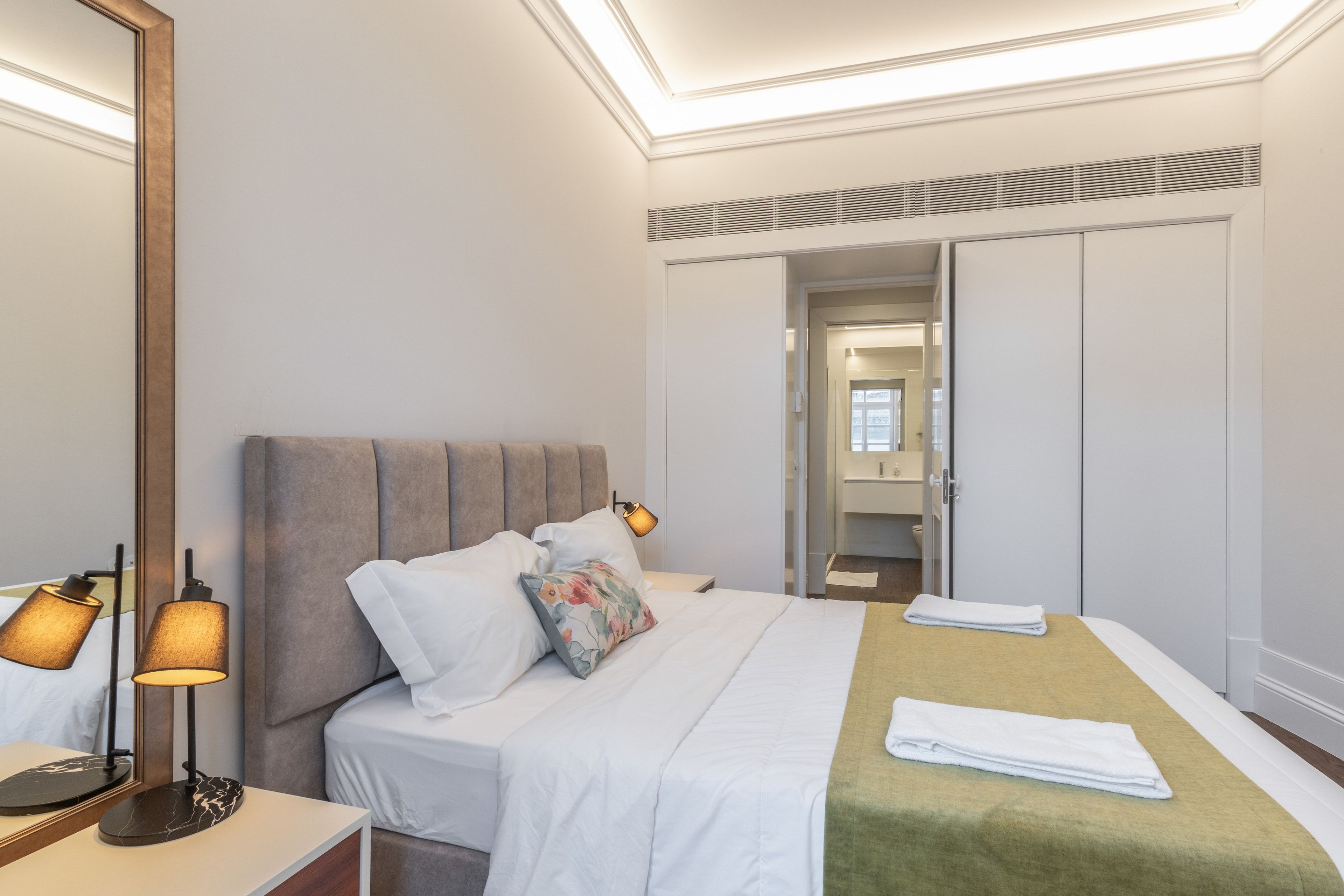 A hotel room with a bed, bedside lamps, a mirror, a wardrobe, and a bathroom visible through an open door.