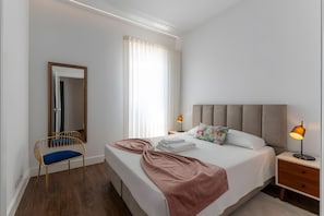 Un dormitorio ordenado con cama, mesitas de noche, una silla, un espejo y una ventana con cortinas.