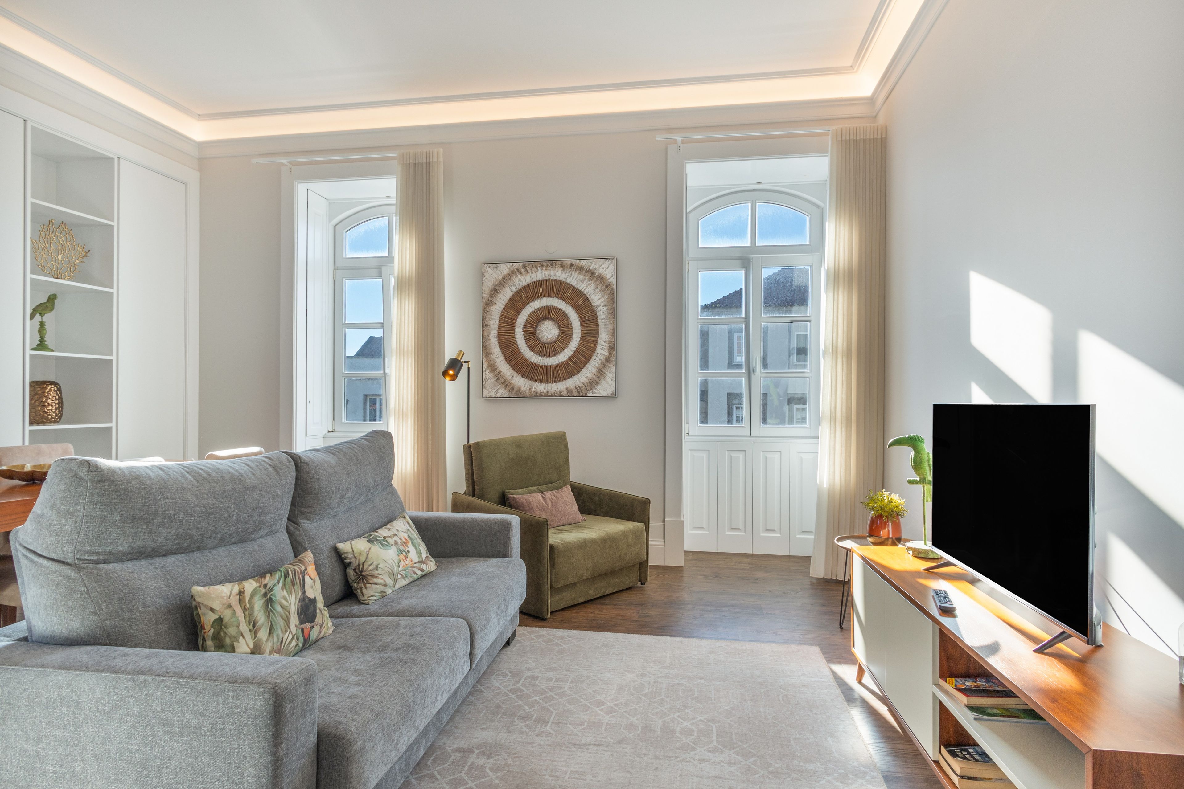 A modern living room with a grey sofa, a green armchair, a large artwork on the wall, and a flat-screen TV on a wooden stand.