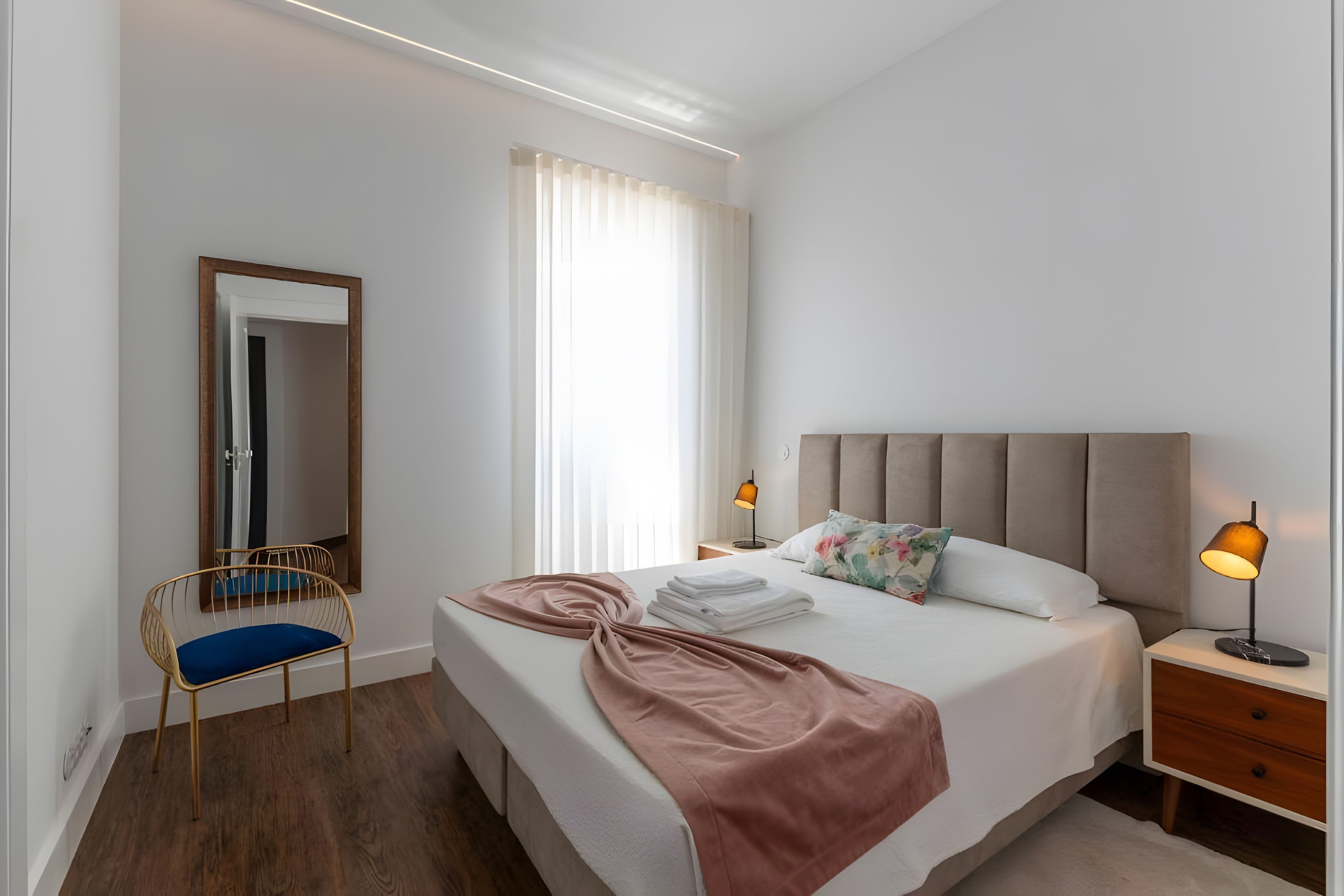 A modern hotel room with a large bed, a bedside table with a lamp, a chair, and a mirror.