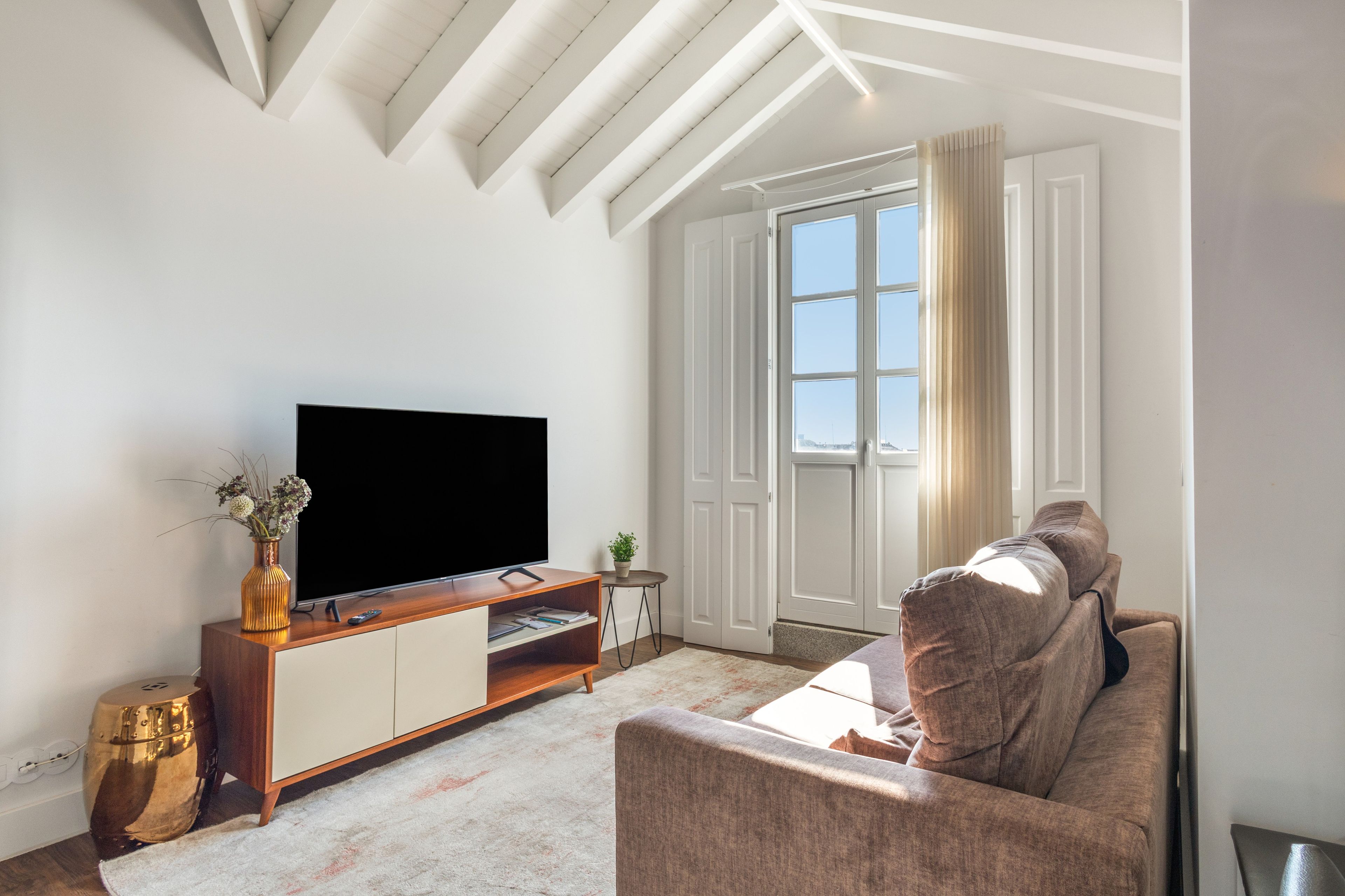 A living room with a flat-screen TV on a wooden TV stand, a sofa, a small side table with a plant, and a large window with curtains.
