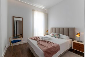 A neatly arranged bedroom with a bed, bedside tables, a chair, a mirror, and a window with curtains. - Apartment 'Guerreiro - T2 2B' (Viana do Castelo)
