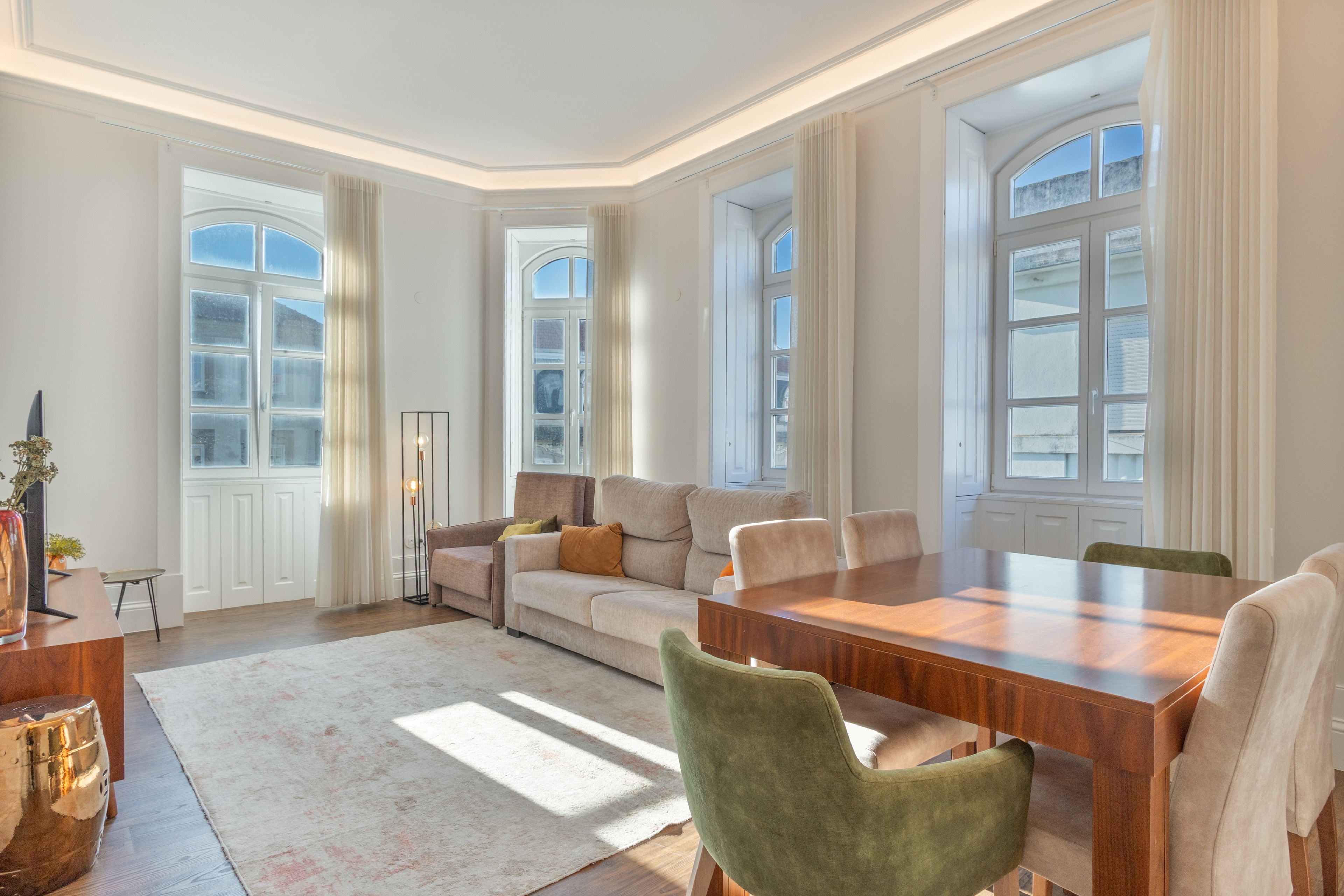 A spacious living room with a wooden dining table, a sofa, and a coffee table. There are large windows with curtains, a floor lamp, and a vase with flowers.