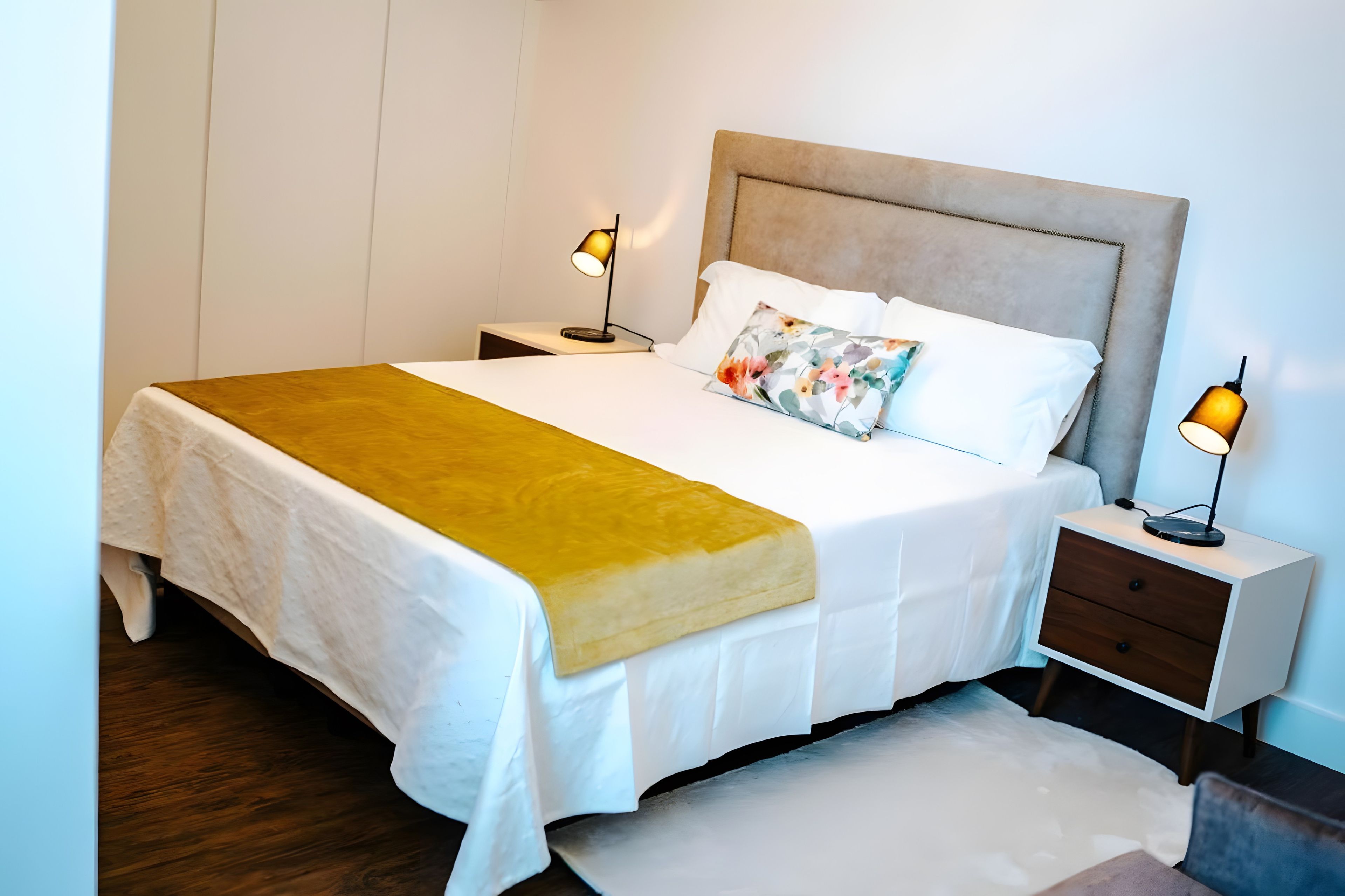 A neatly made bed with a white comforter and a yellow throw blanket, flanked by two bedside tables with lamps, in a room with a wooden floor.