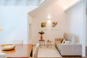 A modern living room with a dining area, featuring a sofa, a table, and two small side tables with decorative items. - Apartment 'Guerreiro - T2 3B' (Viana do Castelo)