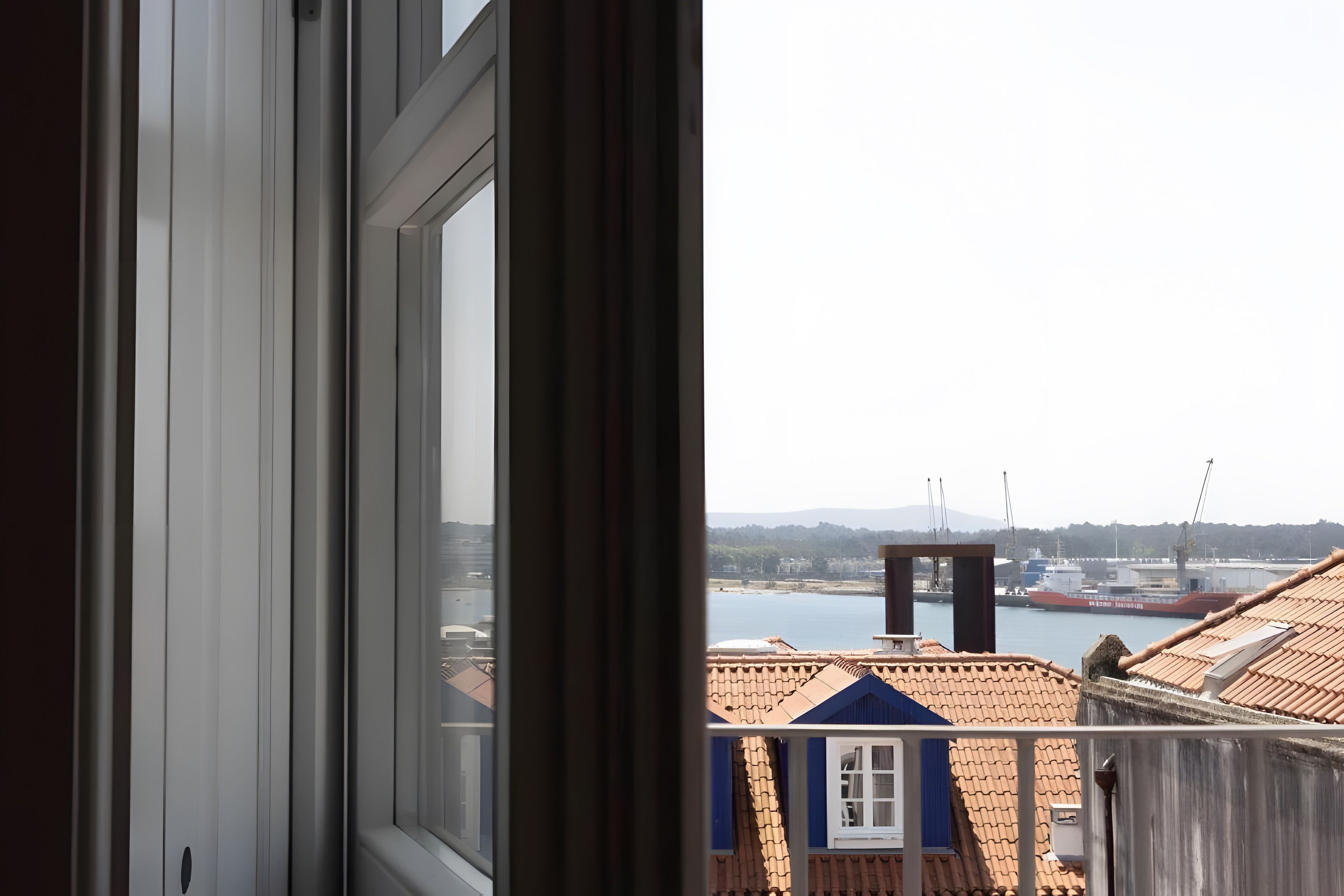 A view through a window with a balcony overlooking a harbor, buildings, and a distant hill.