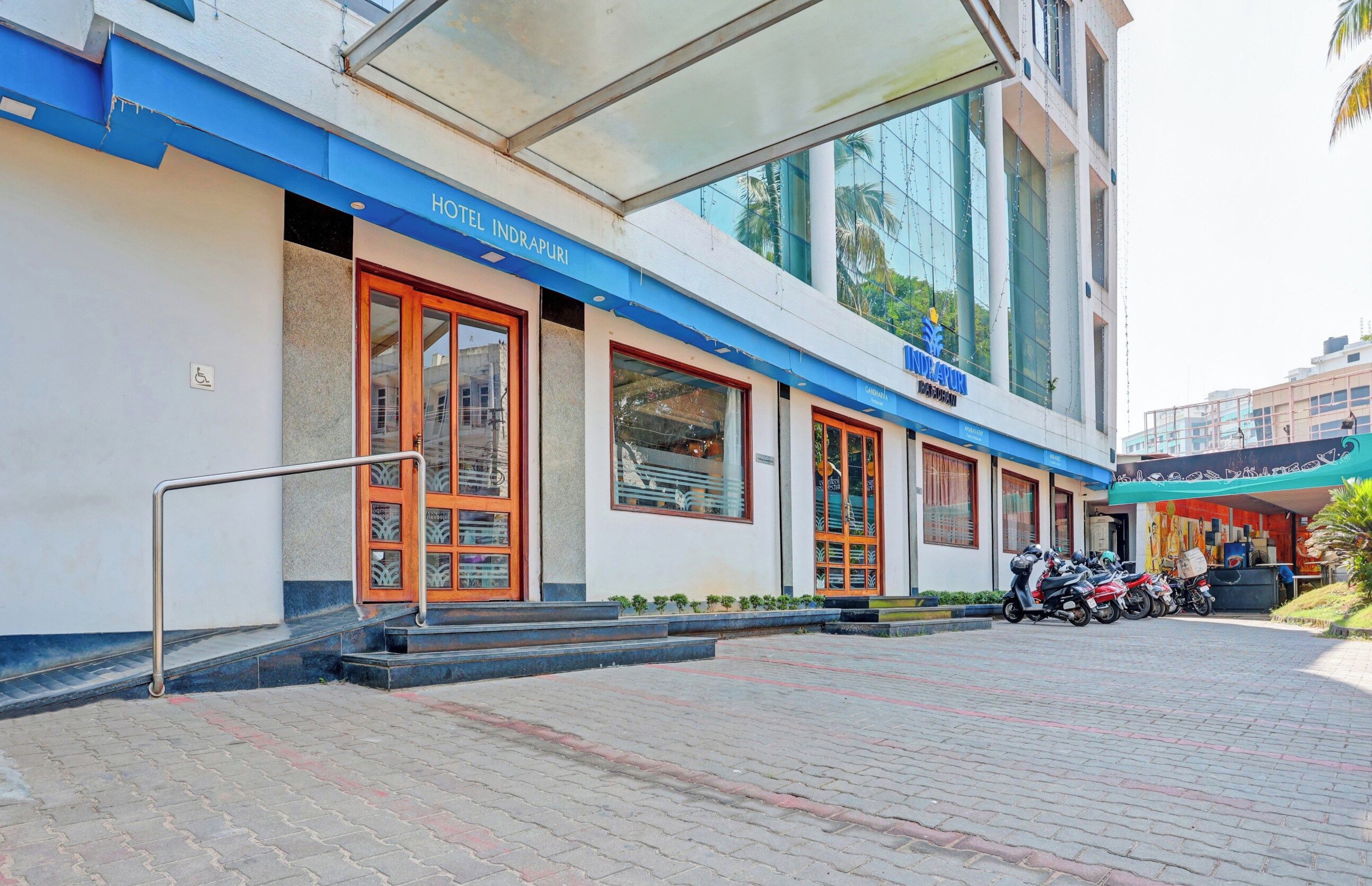 A hotel entrance with a ramp leading to the door, a covered waiting area, and a parking area with scooters.