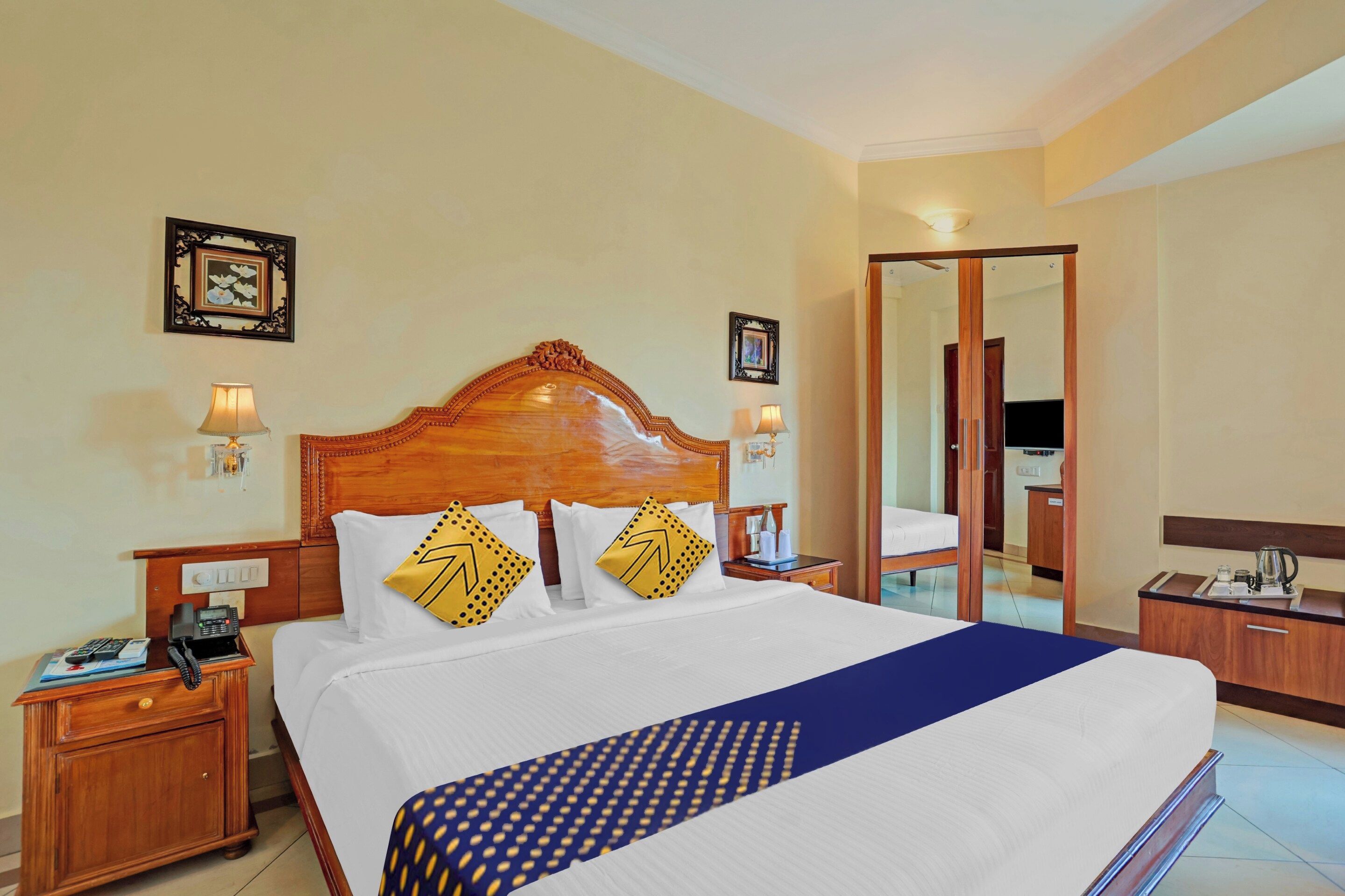 A hotel room with a large bed, wooden headboard, two bedside tables with lamps, a desk with a telephone, and a mirror.