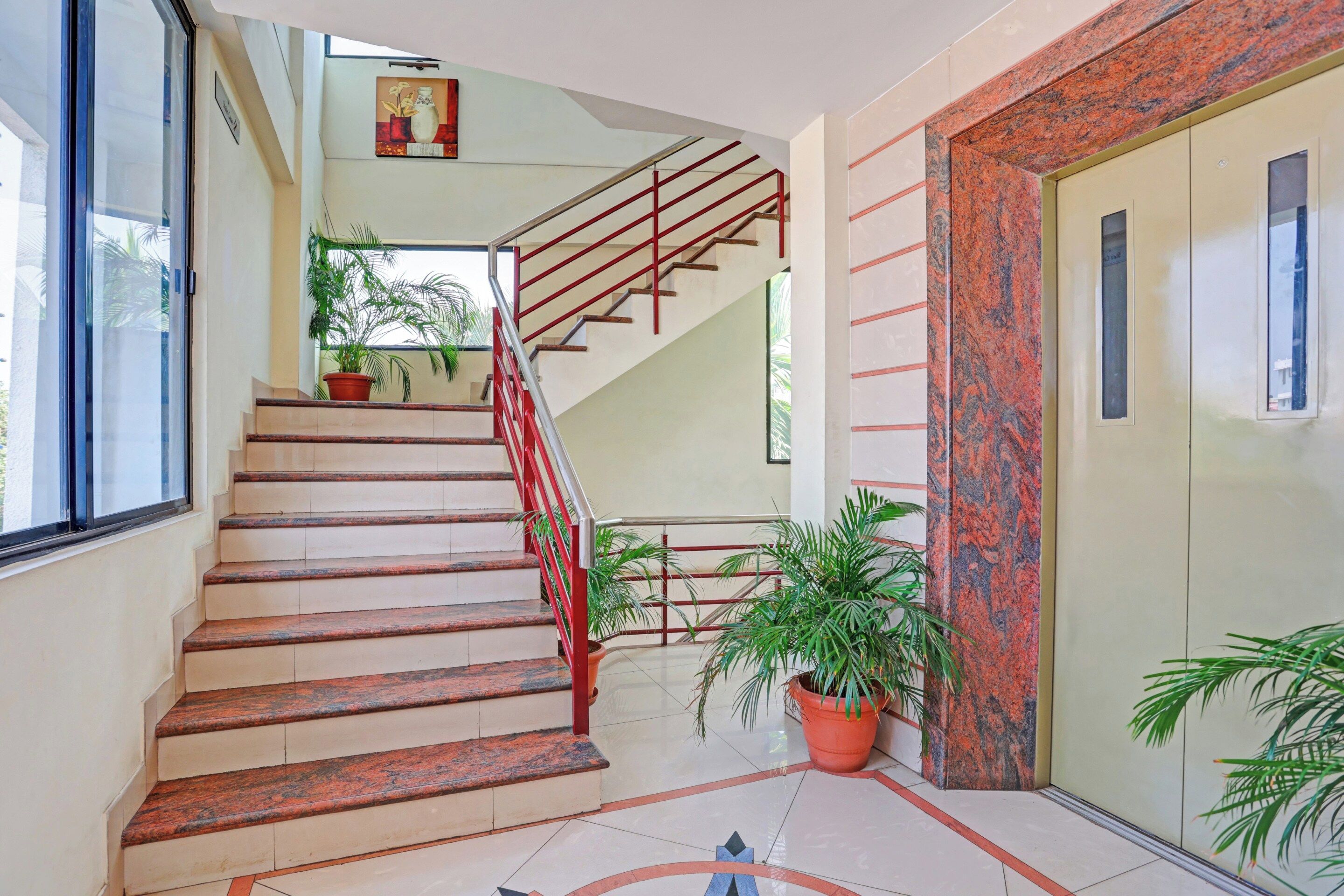 Una escalera con barandales rojos que lleva a un piso superior, rodeada de plantas en macetas y un suelo de baldosas.