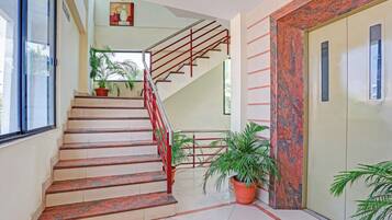 Una escalera con barandales rojos que lleva a un piso superior, rodeada de plantas en macetas y un suelo de baldosas.