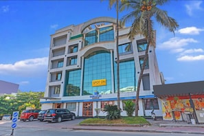 Un hotel de varios pisos con fachada de vidrio moderna, un letrero que indica que es un hotel económico y práctico, y un estacionamiento con varios automóviles.