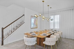 A dining area with a long wooden table set for six, white upholstered chairs, a modern chandelier, and a staircase with white railings. - Magical 9BR Retreat w/ Pool & Game Rooms (Kissimmee)