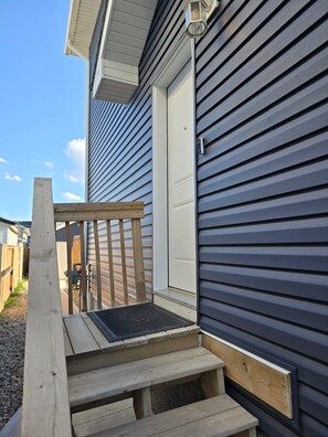 Une terrasse en bois avec un banc et une porte donnant sur une maison dont le revêtement extérieur est de couleur sombre.
