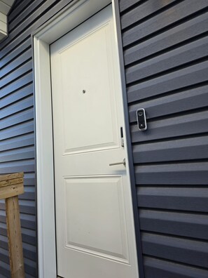Une porte blanche avec un petit judas et un bouton de sonnette, le tout sur un mur recouvert de bardage gris.