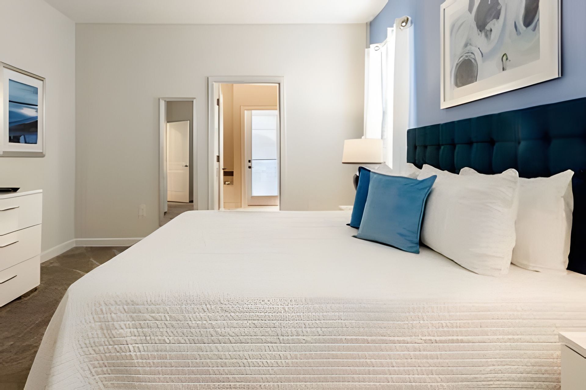 Een hotelkamer met een groot bed, een dressoir, een deur die naar een andere kamer leidt, en een ingelijste foto aan de muur.