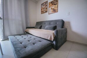 A room with a grey sofa bed, a small table, and a window with curtains. - IBIZA Ap. 109 Hernan (Ipojuca)