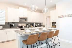 A modern kitchen with white cabinets, a central island with bar stools, a microwave, and a coffee machine. - Magical 5BR Home / Pool & Themed Rooms Near Disney (Davenport)