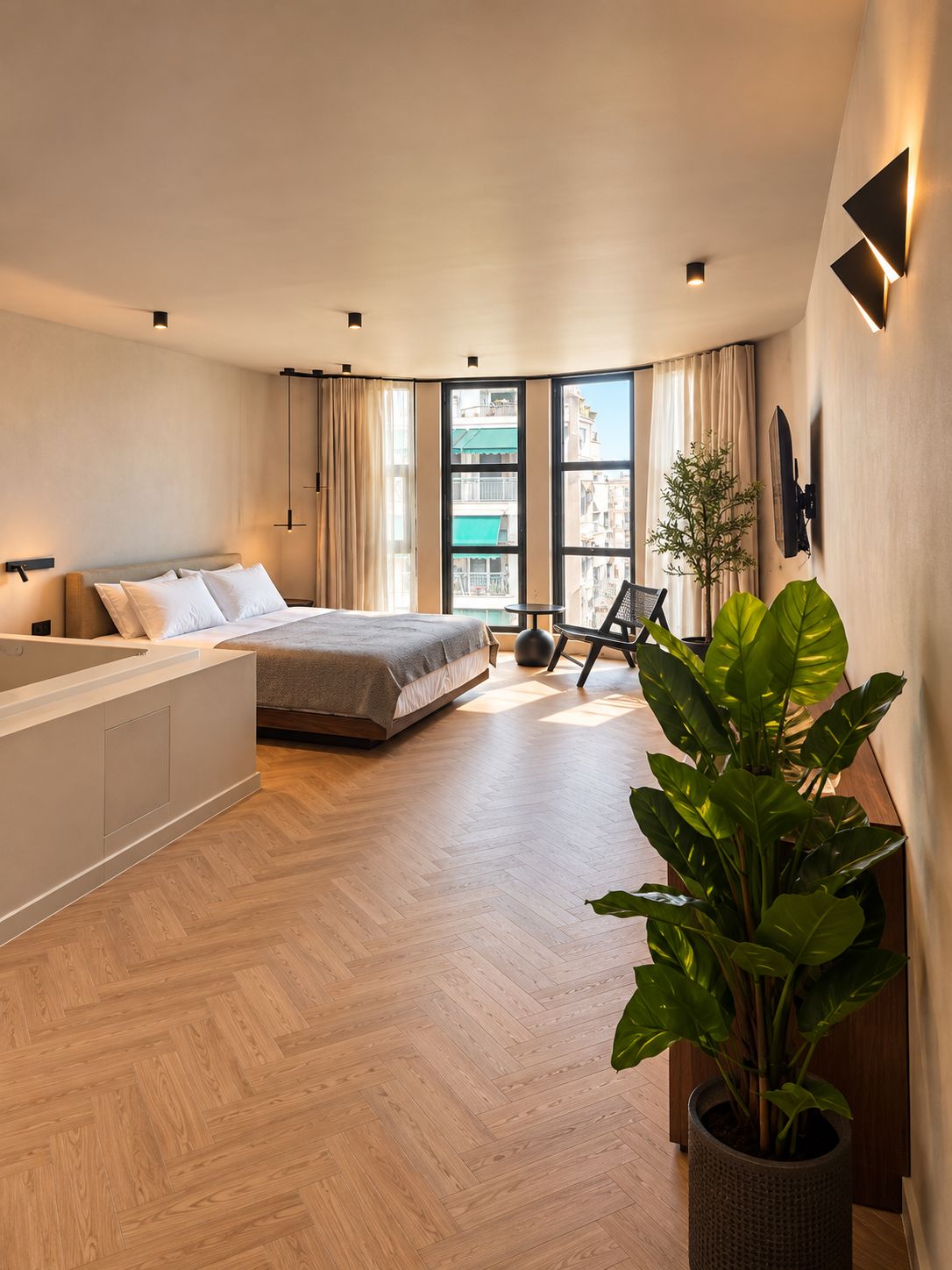A modern bedroom with a large bed, a separate seating area, a view of the city, and a potted plant.