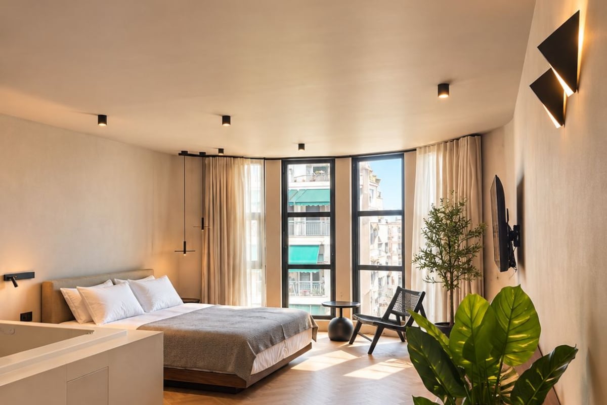 A modern bedroom with a large bed, a separate seating area, a view of the city, and a potted plant.