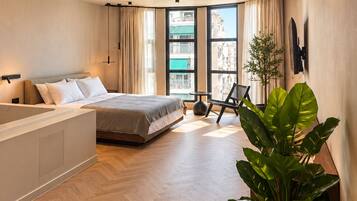 A modern bedroom with a large bed, a separate seating area, a view of the city, and a potted plant.