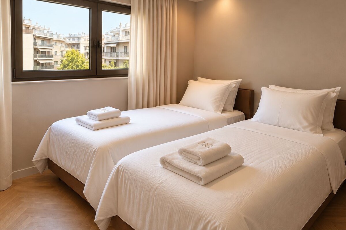 A hotel room with two beds, each with a neatly folded white towel, a window with curtains, and a round table.