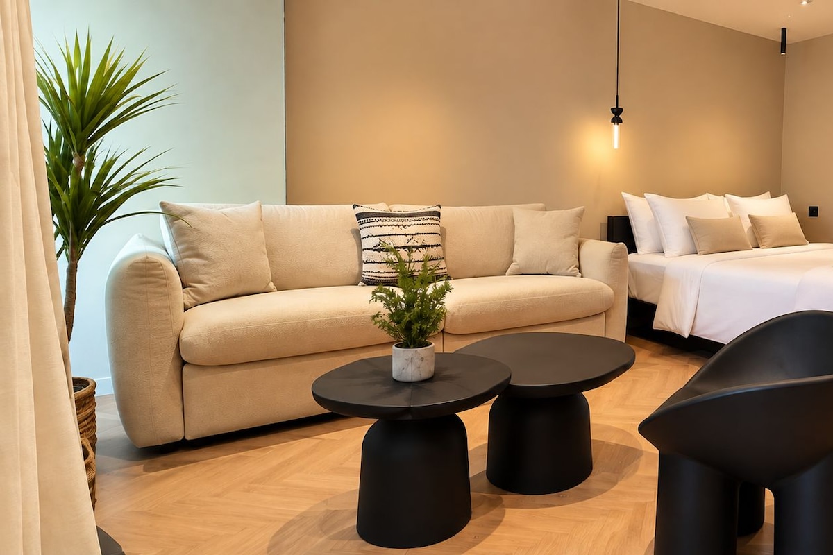 A modern bedroom with a sofa, a bed, two black side tables, and a potted plant.