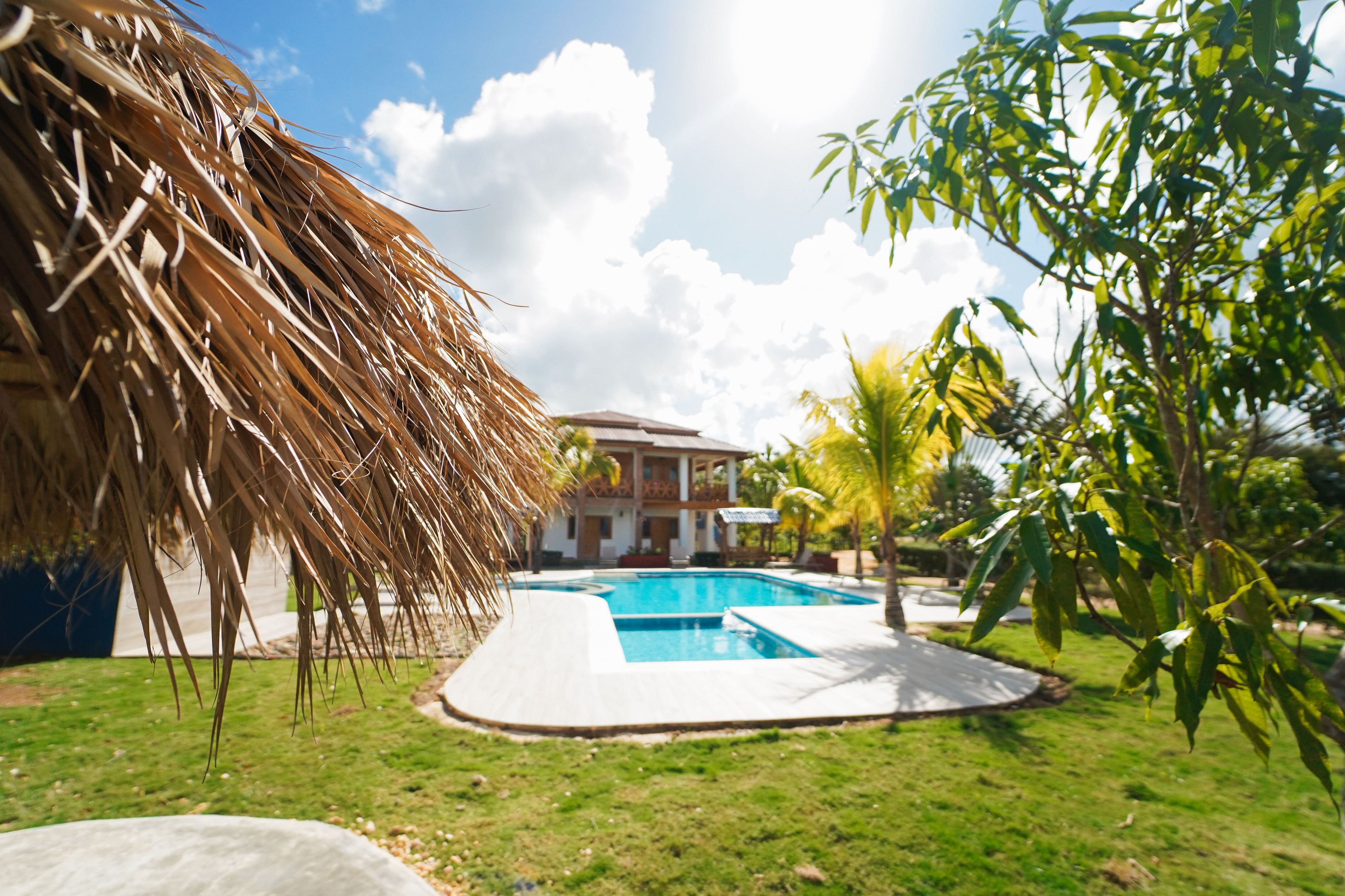 A swimming pool surrounded by grass, a thatched structure, and a building with a balcony.