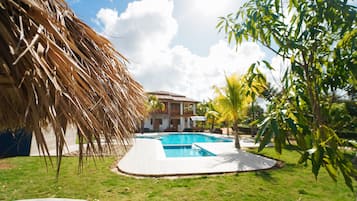 A swimming pool surrounded by grass, a thatched structure, and a building with a balcony.