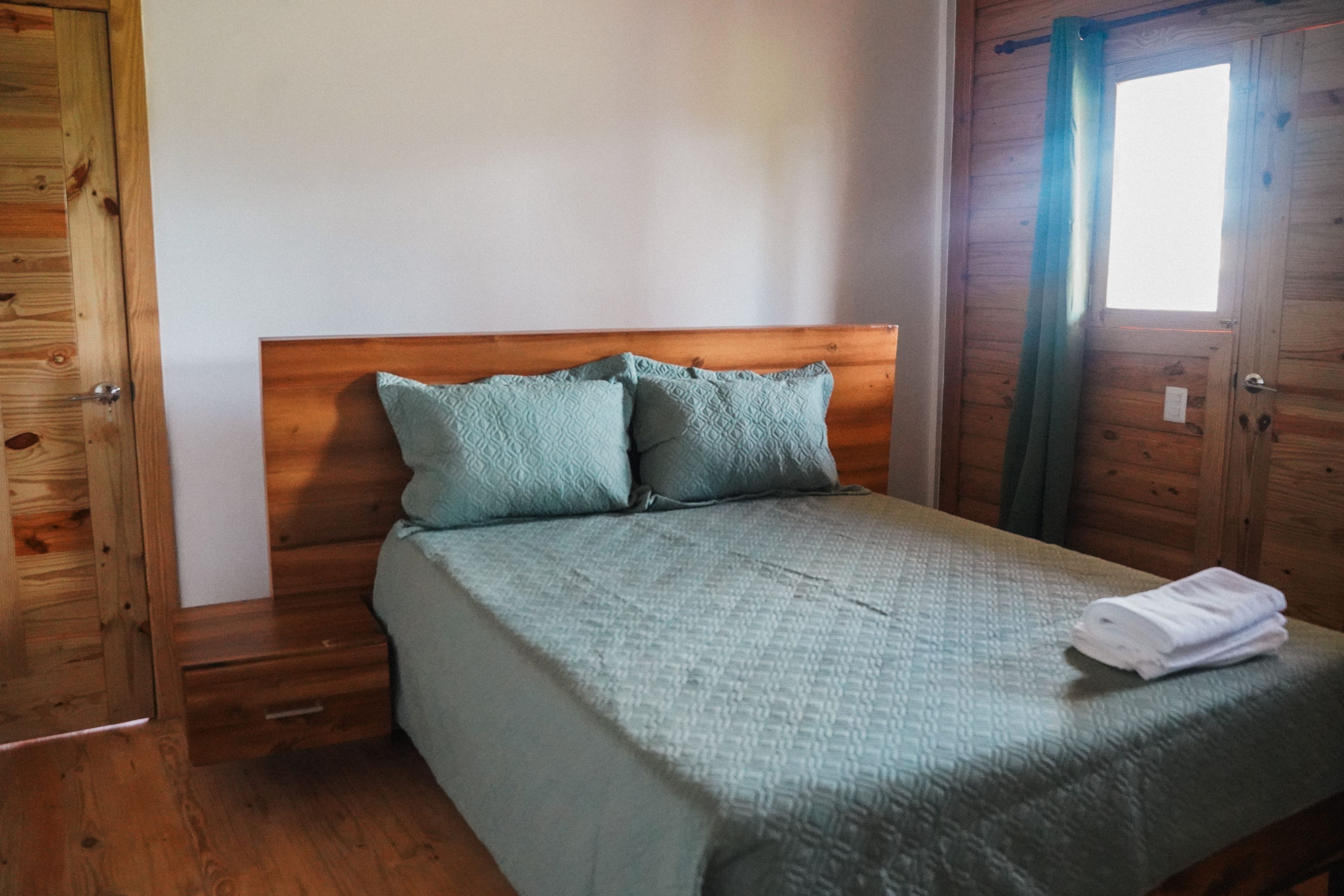 A wooden-framed room with a bed, two pillows, a folded towel, and a window with curtains.