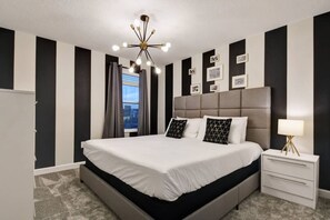 A modern bedroom with a large bed, a bedside table, a nightstand, a chandelier, and a window with curtains. - 9BR Luxury Villa Storey Lake Orlando (Kissimmee)